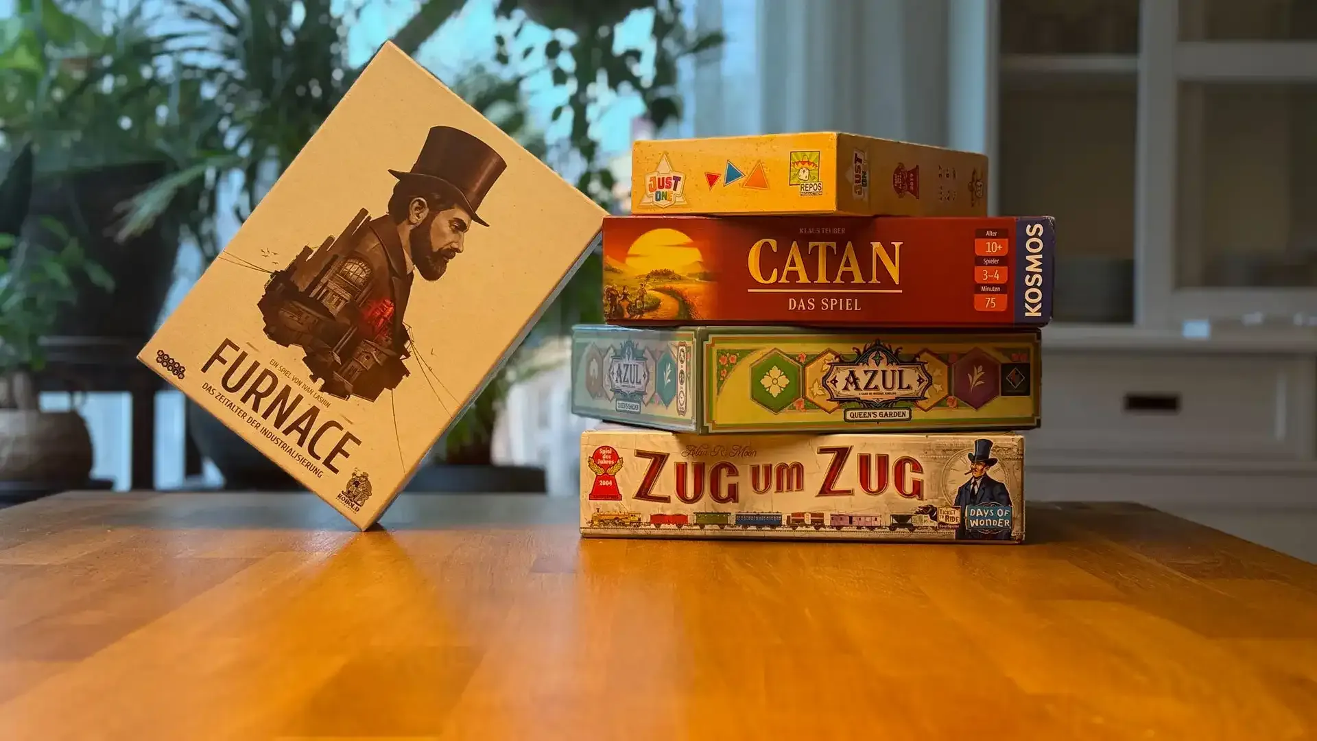 Auf dem Bild ist ein Holztisch. Im Hintergrund sind Pflanzen zu sehen. Auf dem Tisch liegen fünf Spiele: “Zug um Zug”, “Azul”, “Siedler von Catan”, “Just One” und “Furnace”. Vier davon sind aufeinandergestapelt, das fünfte lehnt von links am Stapel.